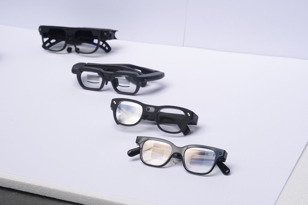 Four pairs of smartglasses lined up on a white surface, from a bulky early frame at the back to slim, ordinary-looking glasses at the front.