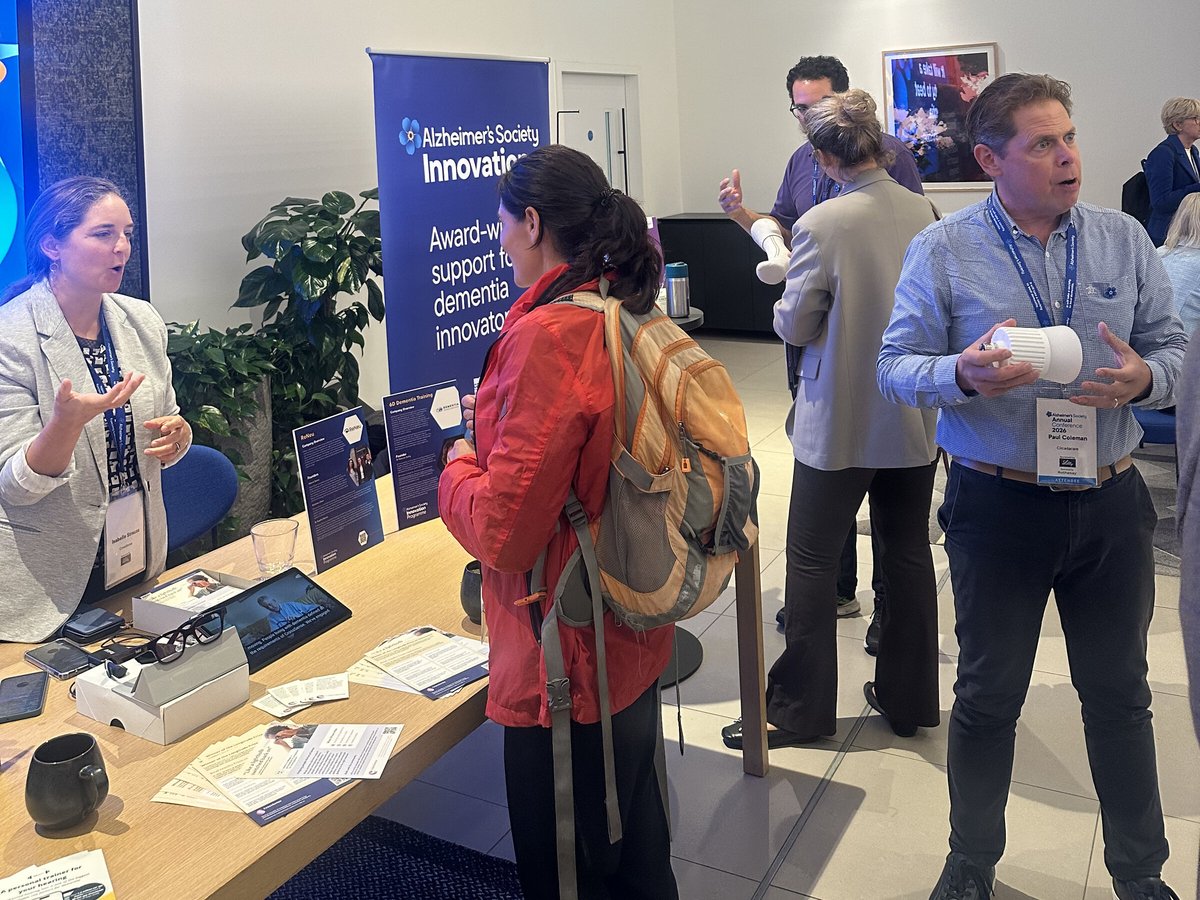 A CrossSense co-founder talks with visitors at a stand with smartglasses and a tablet, beside an Alzheimer's Society Innovation banner.
