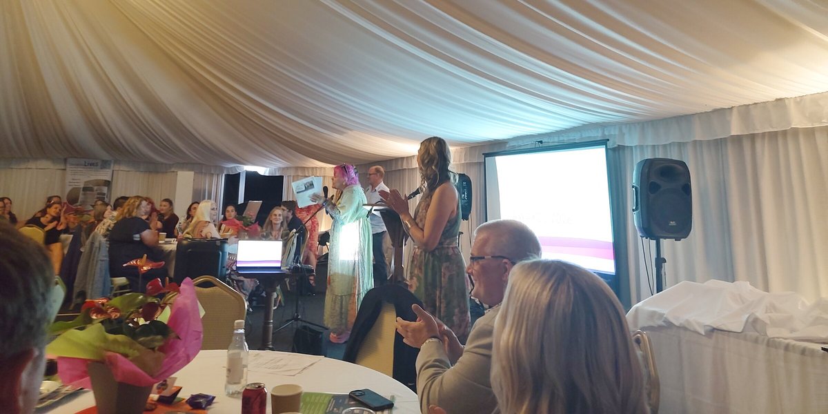 Guests seated at round tables in a marquee watch an award being presented on stage.